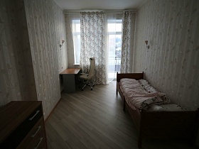 комод, односпальная кровать, письменный стол и компьютерный стул у окна с балконной дверью на открытый балкон двухэтажного кирпичного дома