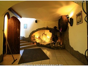 скульптурная композиция старины Мюллера, часть деревянной пивной бочки, яркий светильник под старину на бежевой стене на входе в зал паба в немецком стиле