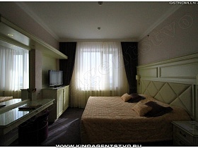 телевизор в углу на тумбе и большое зеркало с гримерным столиком напротив широкой кровати в спальне красивого отеля