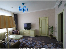 тумба под телевизором, журнальный столик с фигурными ножками и дверь молочного цвета в гостиничном номере  с цветным ковровым покрытием