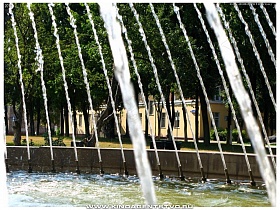 зелень деревьев сквера сквозь равномерные струи воды фонтана в Балашихе