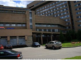 общий вид современного корпуса большой больницы  с машинами на ее территории и разбитыи клумбами