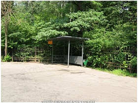 серая автобусная остановка с крышей и скамейкой у металлического забора в лесу
