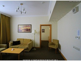 белый торшер и телефон на деревянном круглом стуле у желтого мягкого дивана в спальной комнате гостиничного номера с  люстрой на потолке