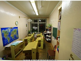 географическая карта, игрушки, папки, различные пособия на открытых полках этажерки, салатневые деревянные парты в светлом учебном кабинете детского центра