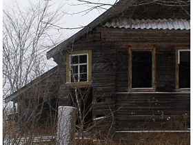 старый неокрашенный деревянный дом с пристройками под треугольной крышей на заросшем травой заснеженном участке