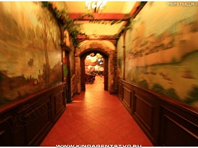  вьющееся комнатное растение под потолком, красочное пано на стенах с деревянными панелями в длинном коридоре  у арочного дверного проема в зал паба в немецком стиле