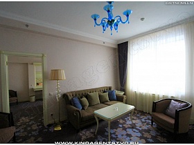 мягкий диван с подушками, кресла и журнальный столик с фигурными ножками  в гостиной с большим окном и голубой люстрой в гостиничном номере