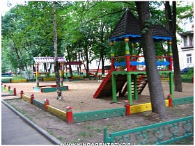  детская площадка с разноцветными игровыми тренажерами за невысоким красочным заборчиком во дворе кирпичных пятиэтажек