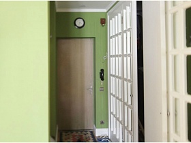 круглые часы над белой входной дверью и коврик на пороге маленькой прихожей с зелеными стенами в двухкомнатной квартире