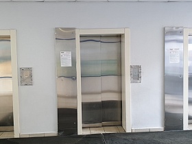 ковровые дорожки у трех лифтов с полированными стальными дверьми в белой стене прозрачного подъезда