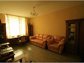 персиковые мягкие диван и кресло, шкафы с посудой в светлой гостиной приличной двухкомнатной квартиры в жилом доме