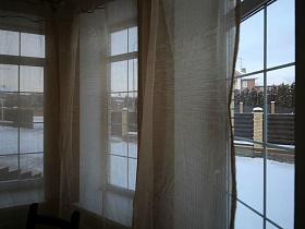 заснеженный двор участка из окон с белыми гардинами и бежевыми шторами кирпичного двухэтажного дома