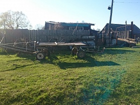 длинная деревянная самодельная телега на автомобильных колесах на зеленой траве у высокого деревянного забора жилого дома старой деревне 2
