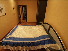 белое с синим стеганное покрывало на большой кровати с серой деревянной спинкой у бежевой однотонной стены спальной комнаты обычной двухкомнатной московской квартиры для съемок кино
