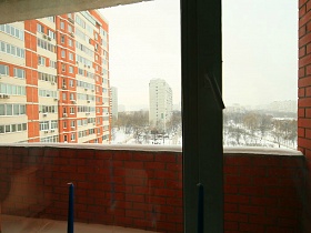 снег на перилах открытого кирпичного балкона через стекло окна и балконной двери трехкомнатной квартиры в переезде (въезде)  молодоженнов