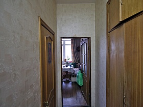 коричневый шкаф для одежды с антресолью у светлой стены длинного коридора с открытой дверью на кухню с обеденным столом просторной семейной трехкомнатной квартиры