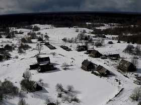 общий вид небольшой заброшенной деревни в снегу в окружении леса с деревянными одинокими жилыми домами