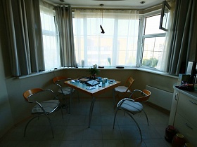 стеклянный  ораньжевый обеденный столик на металлических ножках и стулья со спинками у полукруглого окна с серыми шторамив светлой кухне трехкомнатной квартиры многоэтажного дома
