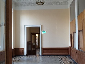круглая люстра с белыми плафонами на высоком белом потолке чистого светлого вестибюля с серой плиткой на полу, деревянными панелями и указателем у входной двери на лестничную клетку