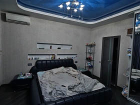 большая черная кровать с мягкой спинкой, светло серым постельным и прикроватными столиками, этажерка в углу в спальной комнате со светло серыми стенами и синим натяжным потолком квартиры студии