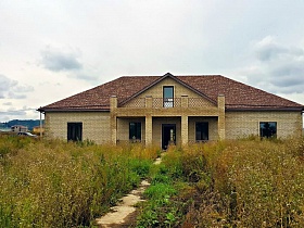 проложенная тропинка среди заросшей травы на участке с недостроенным домом в коттеджном поселке на берегу реки