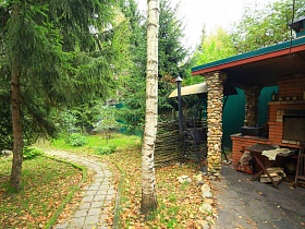 деревянный столик и складной стул у печки под навесом с колоннами из камня на участке с опавшими листьми семейного дома в глухом лесу
