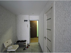 вид на входную дверь соседней квартиры напротив из открытой двери прихожей с обувью на открытой полочке и белым стулом у светло серых стен однокомнатной квартиры