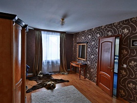 гримерный столик с зеркалом на коричневых цветочных обоях с вензелями на стене спальной комнаты с ковром и медвежьей шкурой на полу современного кирпичного дома