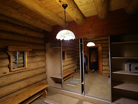 металлический сейф на открытой полке шкафа-купе с зеркальными дверцами, зеркало в деревянной рамке над длинной деревянной скамейкой в  прихожей с белым плафоном подвесной люстры