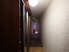 темно-коричневый шкаф-купе с зеркальной дверцей в светлом длинном коридоре с цветным ковром на полу трехкомнатной типовой квартиры