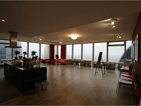 общий вид просторного светлого зала художественной студии с островком кухни, зоной отдыха у панорамных окон и стульями у кирпичной стены с картинами для съемок кино
