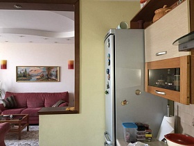 бардовые бра, большая картина над бардовым мягким диваном с подушками у светлой стены студии из кухни с бежевой мебельной стенкой, серым двухкамерным холодильником у стены