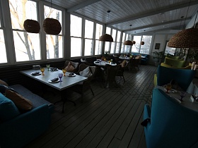 мягкие подушки на диване, плетенных креслах , цветных креслах с высокой спинкой у сервированных столиков в светлой зонированной комнате с деревянным белым потолком и окнами на всю стену стильного ресторана