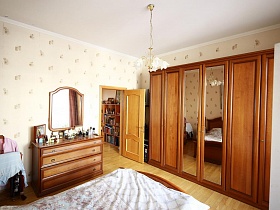 зеркало в деревянной рамке над комодом с туалетными принадлежностями,большой шкаф для одежды с зеркальными дверцами по центру и кровать в светлой спальной комнате простой семейной квартиры в Марьино