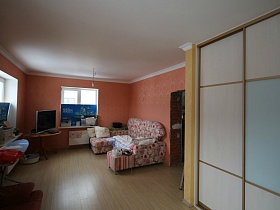 гладильная доска, плоский телевизор на круглом столике, угловой диван с цветочным рисунком, шкаф-купе с матовыми дверцами в комнате с яркими стенами на втором этаже
