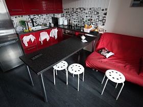 круглые белые стулья у черного обеденного стола на металлических ножках, вишневого дивана на темном полу кухни с вишневой мебельной стенкой и натяжным потолком
