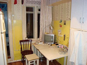 настольная лампа и небольшой серебристый телевизор на углу обеденного стола, табурет и стул со спинкой у высокого окна, белый шкаф и холодильник на кухне с желтыми панелями стен и полосатыми обоями бабушкиной квартиры
