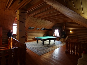 кресло качалка и бильярдный стол на большом светлом ковре напротив окна в комнате под крышей с деревянными балками дома из сруба