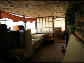 листы крагиса у деревянной стены-сруба,стол, покрытый клеенкой, кресло, хлебница на старом белом холодильнике на деревянном полу застекленной веранды дома в деревне