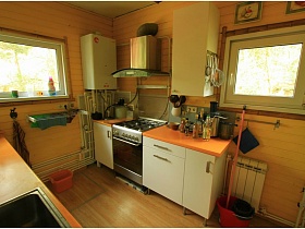 металлическая вытяжка над газовой печкой между шкафами белой мебельной стенки с рыжей столешницей в кухне с колонкой на стене