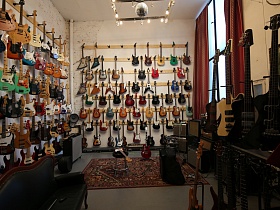 огромный выбор разнообразных электро и электро акустических гитар в светлой комнате с большим окном, диваном для посетителей и цветным ковром на полу стильного музыкального магазина