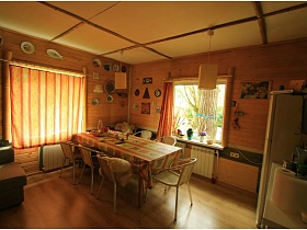 плетенные кресла вокруг длинного обеденного стола, белый холодильник в гостиной с квадратными плафонами на светлом потолке и короткими полосатыми шторами на больших окнах