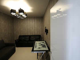 два дивана, черный столик на ножках на полу, люстра с квадратными черными плафонами на светлом потолке  небольшой гостевой комнаты через открытую белую дверь