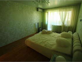 большая белая кровать в светлой спальне с салатневыми гардинами на окне простой двушки