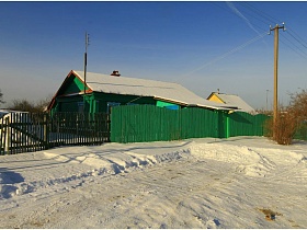 заснеженная крыша жилого деревянного дома, окрашенного в зеленый цвет за высоким зеленым деревянным забором в зимнее время
