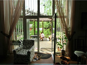 мягкое кресло и высокий комнатный цветок у стены с панорамными окнами и стеклянной дверью с видом на беседку под крышей в саду современного дома с балконом и бассейном