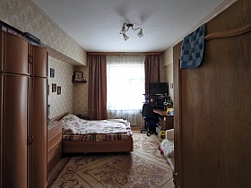 шкаф для одежды, комод и большая деревянная кровать в спальной комнате с коричневым ковром на полу и коричневыми шторами на окне просторной семейной трехкомнатной классической квартиры в сталинском доме