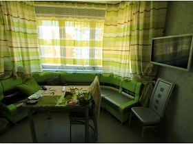 полукруглый мягкий уголок в зеленом цвете с обеденным столом у окна кухни с зелеными шторами в двухкомнатной квартире жилого дома в новострое