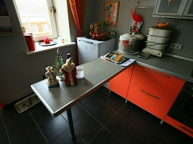 электроприборы на серой столешнице ораньжевой кухни, ораньжевая картина над морозильной камерой у окна с ораньжевой гардиной семейной трехкомнатной квартиры